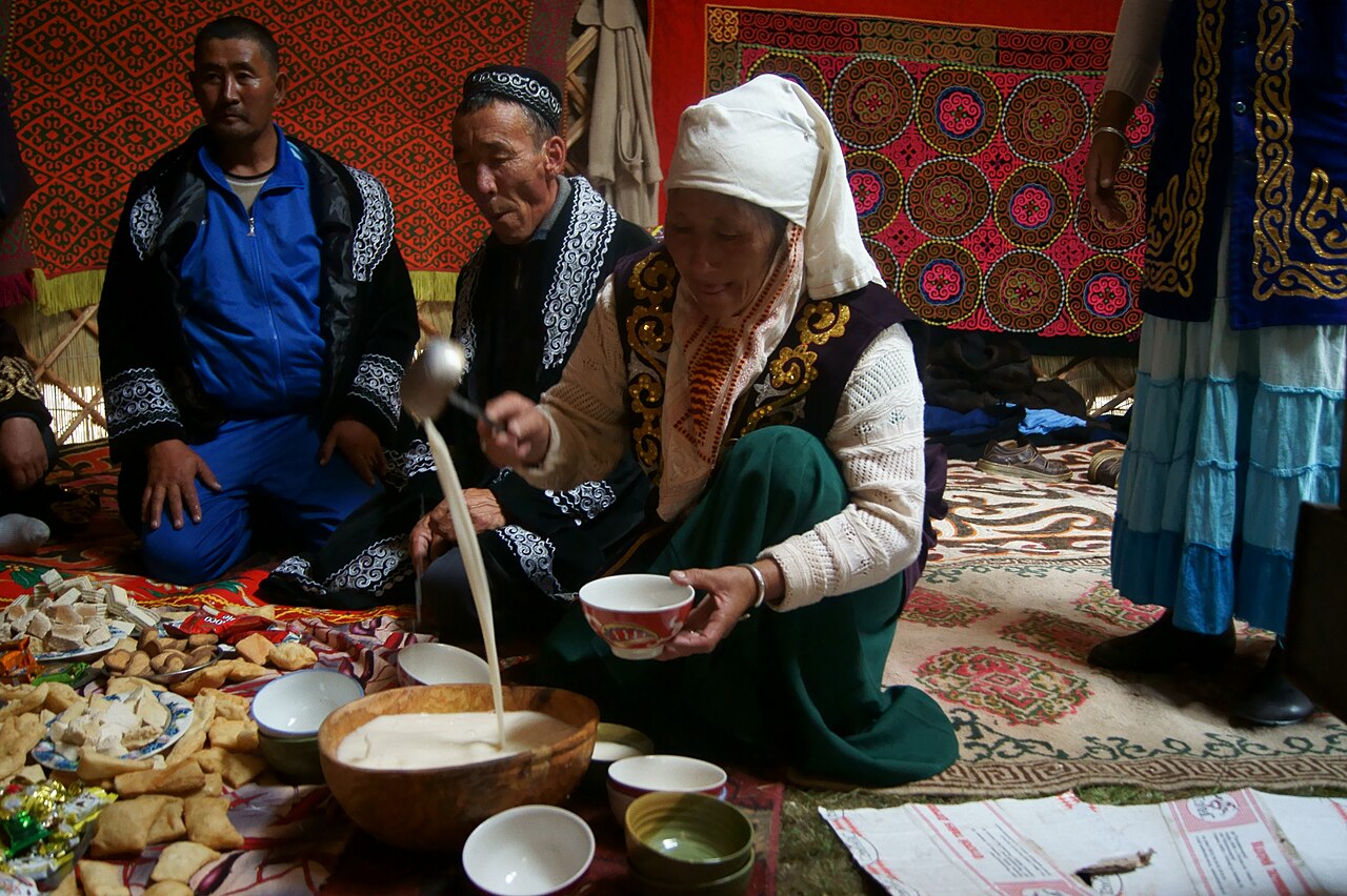 A Kazakh woman pouring tea into a piala at a dastarkhan spread with baursak and sweets